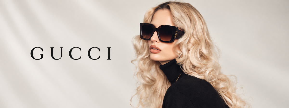 A blonde model wearing oversized dark Gucci sunglasses and a black turtleneck, posed against a light draped fabric backdrop with the GUCCI logo printed on the left.